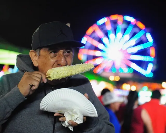 Featured Photos 10-18-2024. Corona resident Hector Manriquez enjoys corn on the cob as the Ferris wheel turns behind him at the St. Edward Catholic Church Carnival in Corona. Credit: Photo by Jerry Soifer