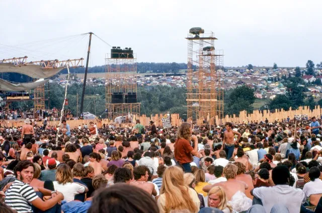 August 15. Joe Cocker performs on stage at left before crowd and huge lighting/sound towers. Cars and tents can be seen in the background. Credit: Woodstock Whisperer
