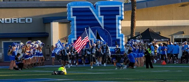 Bearing the American Flag, Norco’s Trevor Schneider leads Norco teammates and the cheer squad onto the home turf, kicking off the 2026 football season versus neighboring rival Eastvale Eleanor Roosevelt.
Credit: Photo by Gary Evans